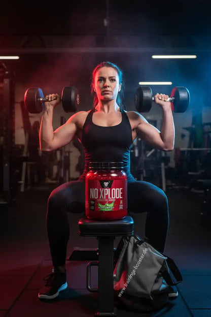 Muskulöse Frau trainiert im Gym mit Hanteln hinter BSN Nutrition N.O. XPLODE Pre Workout Booster.